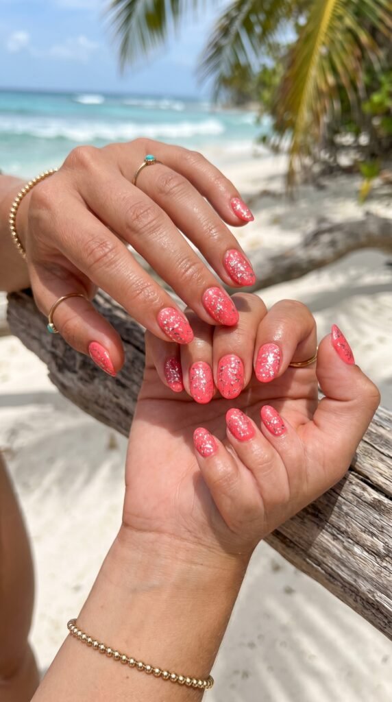 Hands with coral base nail polish featuring scattered silver glitter sparkle creating tropical beach-inspired summer manicure with vibrant playful aesthetic
