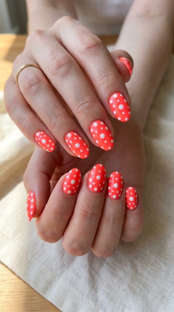 Hands with neon coral nail polish base featuring crisp white polka dots in retro classic pattern creating playful vintage-meets-modern design for cheerful summer manicure






